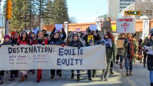 Demonstrators march in the street behind a banner that says "Destination: Equity" on a sunny day in Moncton.