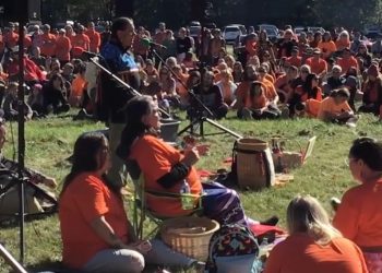 A large group of people, many dressed in orange is gathered outdoors. People appear to be sitting in a circle on the grass.