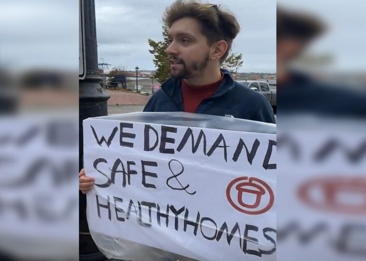 Someone with brown hair and a light beard holds a banner that says "WE DEMAND SAFE & HEALTHY HOMES" against a grey sky.
