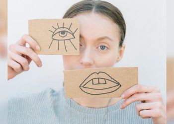 Une femme tient deux pièces de papiers devant son visage. Un oeil et une bouche sont dessinés sur le papier