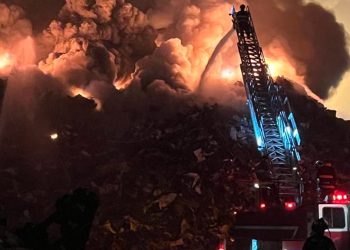 Smoke billows from a scrapyard; a ladder truck is visible hosing down the burning scrap.