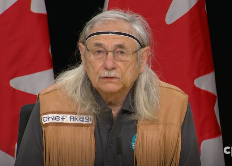 Peskotomuhkati Chief Hugh Akagi is wearing a leather vest with a name tag that reads "Chief Akagi," and stands in front of a backdrop featuring Canadian flags.