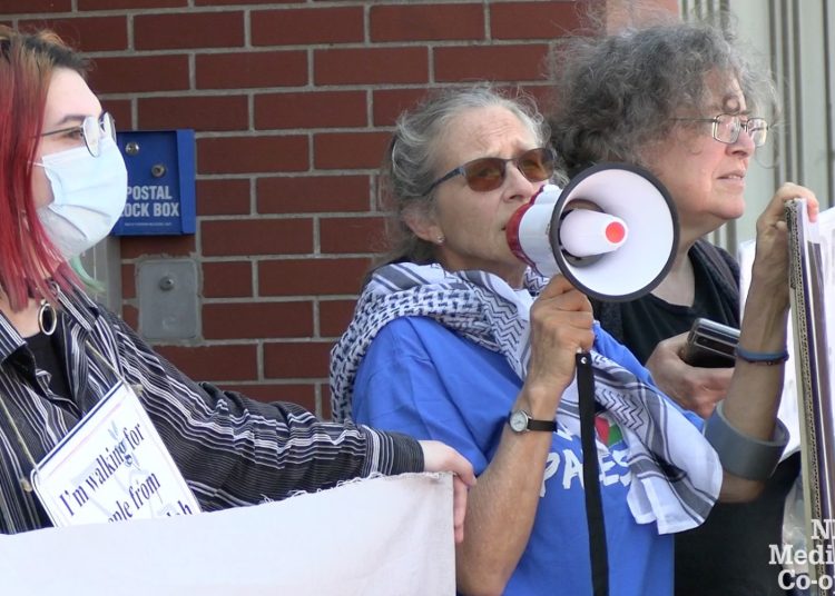 Three protesters are pictured standing outside a brick building, holding a banner, a megaphone and a placard.