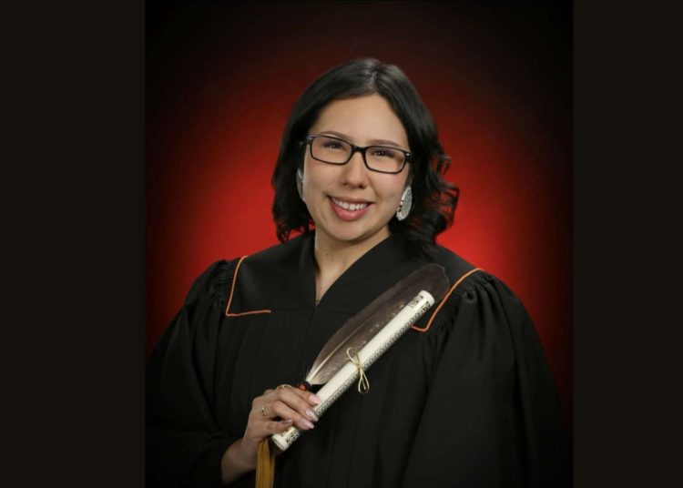 Zoey Milliea of Elsipogtog First Nation, 2024 graduate, holding a diploma with a feather. Wearing cap and gown, red and black background.