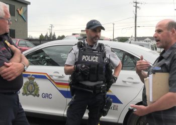 A man wearing a plaid shirt holding a folder of documents under his arm speaks to a uniformed police officer and a uniformed fire marshal in front of an RCMP cruiser.