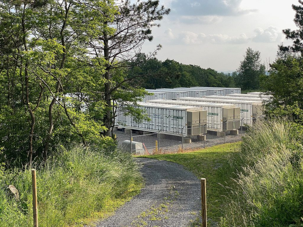 Rows of large white battery storage containers in a fenced facility surrounded by trees and greenery.