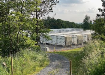 Rows of large white battery storage containers in a fenced facility surrounded by trees and greenery.