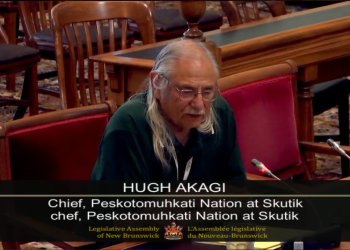 Hugh Akagi, Chief of the Peskotomuhkati Nation at Skutik, speaks into a microphone at the Legislative Assembly of New Brunswick. He has long gray hair and glasses, and is wearing a dark shirt. The chamber is furnished with wooden chairs and desks, and the caption on screen identifies him by name and title.