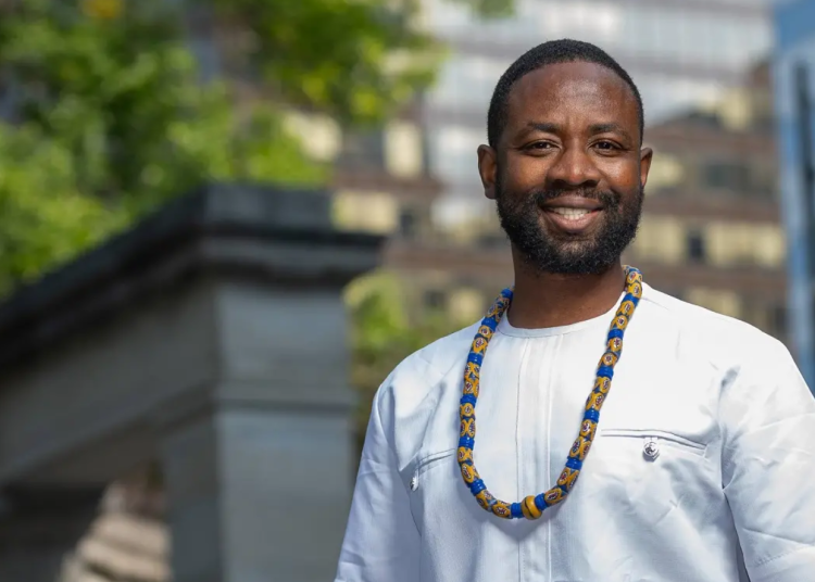 Wendell Nii Laryea Adjetey, smiling outdoors, wearing a white shirt and a beaded necklace, with blurred buildings and greenery in the background.