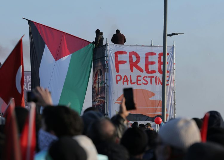 Large crowd of protesters holding Palestinian flags in front of a banner that reads “Free Palestine,” with two people standing on a scaffold above the crowd.