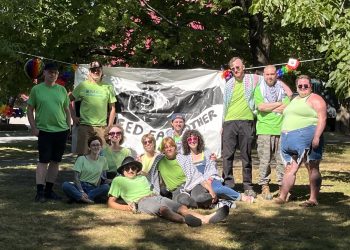 A group of about 15 organizers and volunteers wearing bright green shirts pose together in Queen Square, Saint John, in front of a large banner reading “Feed Each Other.” Trees and decorations are visible in the background.