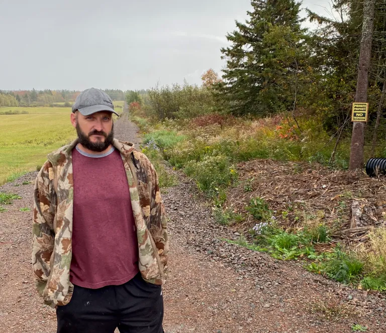Cory Estabrooks stands on a gravel road wearing a camouflage jacket, maroon shirt, and cap. A “Private Property No Trespassing” sign is visible on a tree to his right, with fields and trees stretching out behind him.