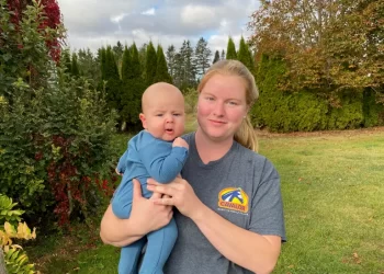 Katie Hess stands outdoors on a grassy lawn holding her 3-month-old son Tatum, both dressed in casual clothes, with trees and shrubs in the background.
