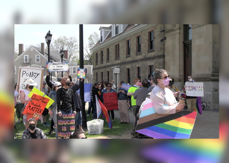A group of people gather outside a stone building for a rally supporting Policy 713, which protects LGBTQ2+ students in New Brunswick schools. Participants hold colorful signs and Pride flags, with messages such as “Queer and Trans Students Matter,” “Policy 713 is about respect,” and “Queer education is non-negotiable.” Many attendees wear masks, and one person in the foreground holds a Pride flag and a coffee cup.
