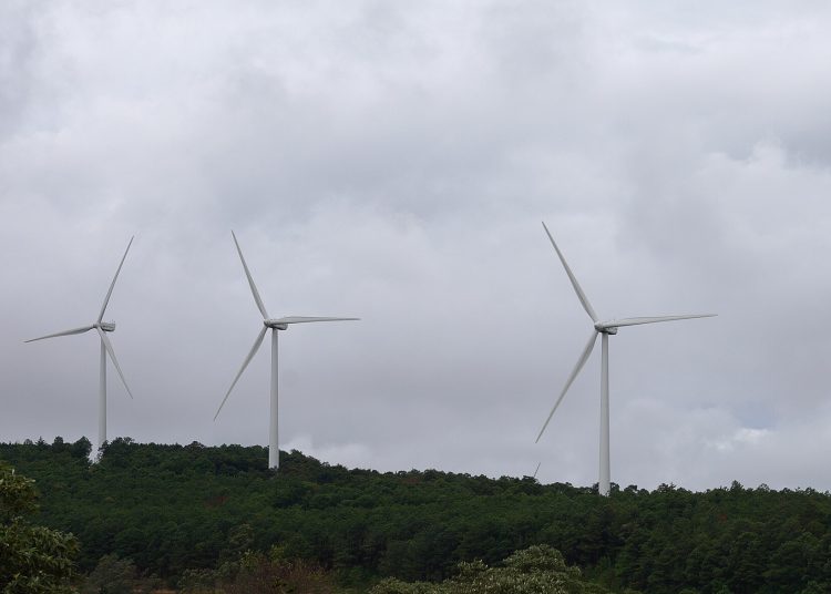 Wind turbine standing among trees with a cloudy sky in the background.