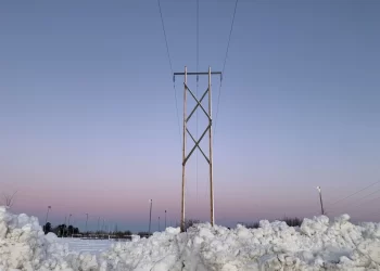 A photograph of a wooden H-frame utility pole supporting three overhead power lines, silhouetted against a pale purple and blue winter sky. Large mounds of snow are piled in the foreground.