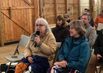 Terry Jones (left), holding a microphone, and Juliette Bulmer (right), sitting side-by-side during the community meeting. They are seated in chairs in a rustic, wooden barn setting.