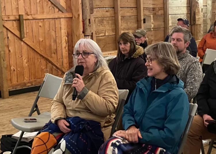 Terry Jones (left), holding a microphone, and Juliette Bulmer (right), sitting side-by-side during the community meeting. They are seated in chairs in a rustic, wooden barn setting.