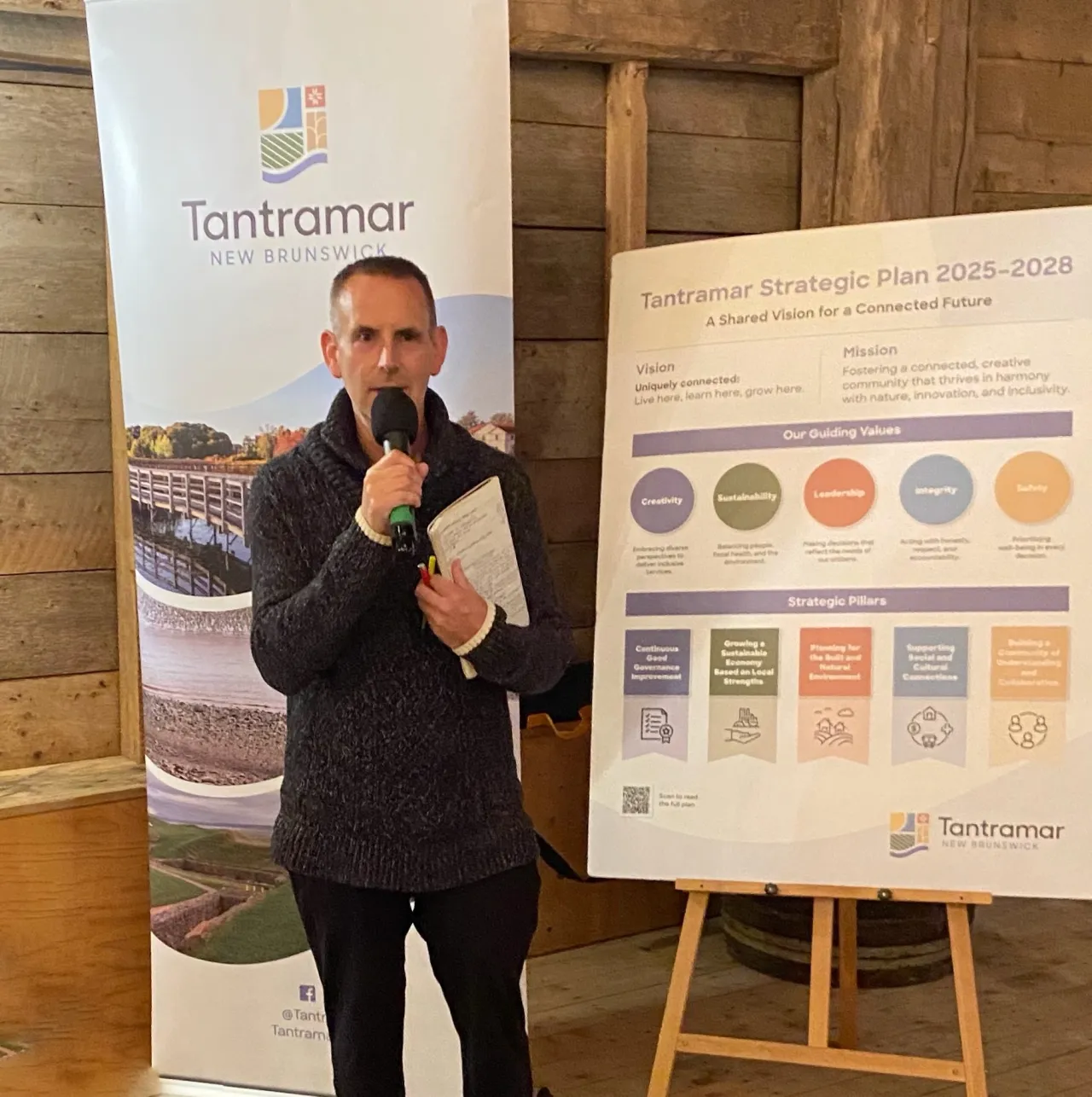 Tantramar Mayor Black, wearing a dark sweater, stands in front of a banner for the Town of Tantramar and a display board showing the "Tantramar Strategic Plan 2025-2028," speaking into a microphone.