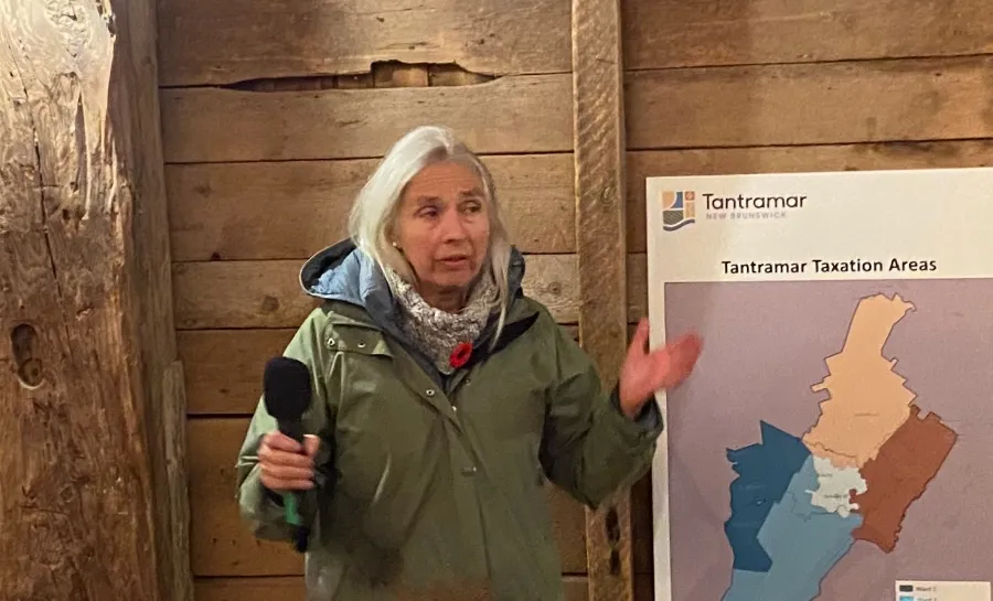 Sackville resident Meredith Fisher, wearing a green jacket, speaks to the audience, gesturing with her hands. A map titled "Tantramar Taxation Areas" is partially visible next to her.