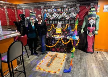 Two women standing next to a colorful Day of the Dead (Día de Muertos) altar in a room decorated for the event.