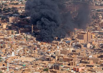 Aerial view of a city in Sudan during the 2023 war, showing large plumes of thick black and grey smoke rising from buildings, illustrating the widespread destruction of urban infrastructure.
