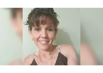 A portrait of Erin Brooks, an Indigenous woman with brown hair, bangs, and a warm smile. She is looking directly at the camera, wearing a dark lace-trimmed top and two thin gold necklaces. The photo is a close-up against a neutral, light-colored background.