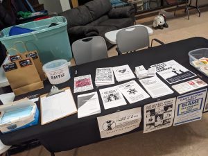 A close-up view of the resource table. Pamphlets with titles like "Mutual Aid," "A Brief History of Capitalism," and "Really Really Free Market" are neatly arranged. A sign on the table reads "This is a community space — Police not welcome."