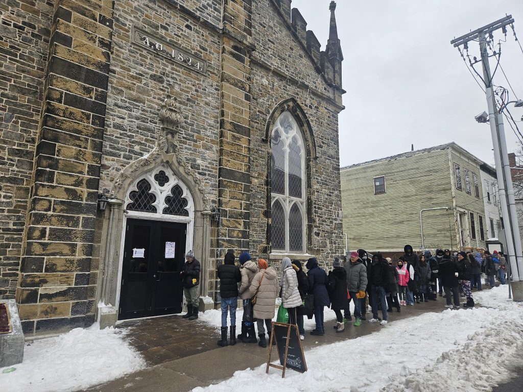 A wide shot of a historic stone church building with a plaque reading "A.D. 1824." A long line of people in heavy winter coats and hats stretches down the snowy sidewalk, waiting to enter through black double doors marked with "Free Store" signs.