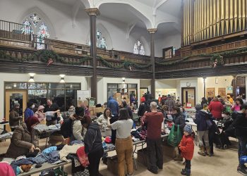 The interior of a large, high-ceilinged church sanctuary. The room is filled with people browsing long tables piled high with clothing and winter gear. A large pipe organ and stained-glass windows are visible in the background, contrasting with the busy, grassroots atmosphere of the clothing swap.
