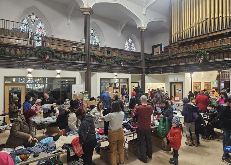 The interior of a large, high-ceilinged church sanctuary. The room is filled with people browsing long tables piled high with clothing and winter gear. A large pipe organ and stained-glass windows are visible in the background, contrasting with the busy, grassroots atmosphere of the clothing swap.