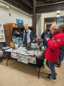 A volunteer in a denim vest stands behind a black table covered in zines and stickers. A woman in a red winter parka browses the materials. Signs on the front of the table read "Can’t afford food? Blame billionaires, not immigrants" and "Autocrats don't create anything."