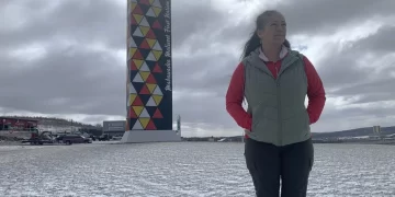 Chief Patricia Bernard of Madawaska First Nation stands in a snow-dusted field, looking toward the horizon with a thoughtful expression. She is wearing a red long-sleeved shirt, a grey quilted vest, and dark trousers with tall boots. In the background, a tall, prominent monument stands against a cloudy sky, featuring colorful geometric patterns in yellow, red, and black, with the words "Madawaska Maliseet First Nation" written vertically along its side.