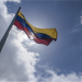 The flag of Venezuela, featuring three equal horizontal stripes of yellow (top), blue (middle), and red (bottom). In the center of the blue stripe, there is an arc of eight white five-pointed stars. The flag is waving behind a blue sky.