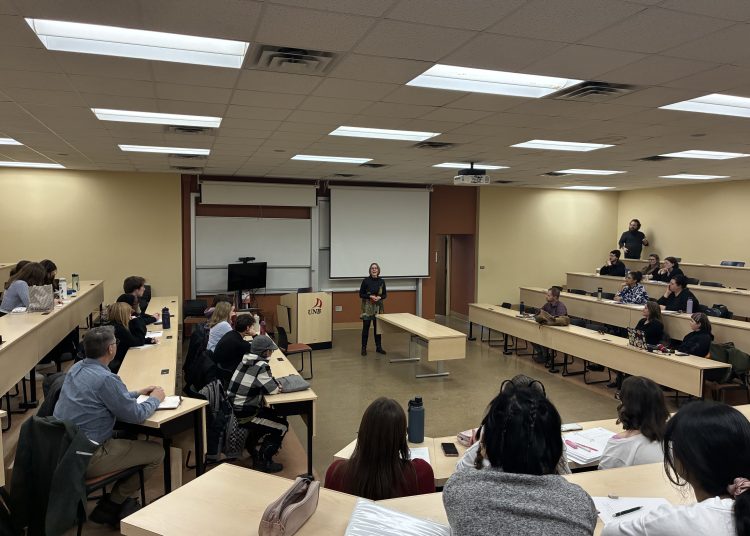 A historian stands in the center of a tiered UNB classroom, leading a discussion with a group of attentive students and faculty seated in a semi-circle.
