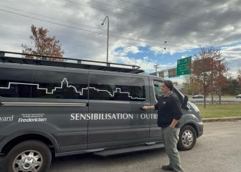 Max Goodine stands beside a grey John Howard Society outreach van on a cloudy day. He has one hand on the door handle and the other in his pocket. The van features a white skyline graphic and the words "SENSIBILISATION | OUTREACH" on the side. A green highway sign and autumn trees are visible in the background.