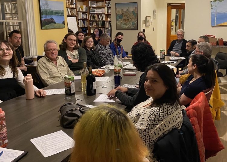 A group of approximately twenty people of diverse ages and backgrounds sit around a long, dark wood table in a brightly lit community room. They are engaged in a meeting, with some taking notes and others listening intently. The table holds papers, water pitchers, and snacks. Art pieces and a climbing green plant decorate the cream and yellow walls in the background.