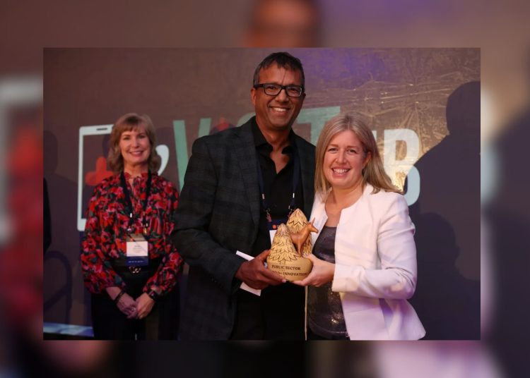 A medium shot of three people at an awards ceremony. On the left, Amy McLeod stands in a red floral blouse. In the center, Dr. Hanif Chatur holds a wooden trophy carved with trees and a deer. On the right, Premier Susan Holt smiles while holding the award with him.