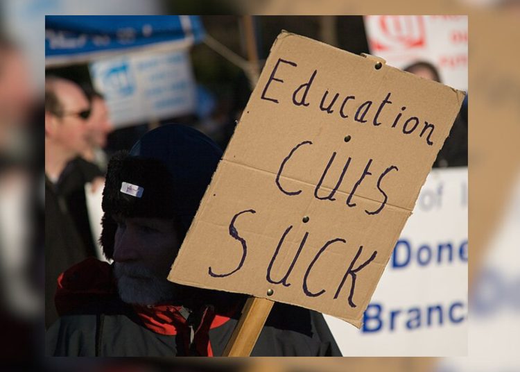 A close-up, medium shot shows a person at an outdoor protest holding a cardboard sign that reads "Education cuts SUCK" in hand-written, blue marker. The person is wearing a dark winter hat with earflaps and a black and red jacket. Other protesters and banners are blurred in the background under bright sunlight.