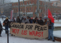 A group of people in winter coats stand in a snowy downtown square in Fredericton, holding a large banner reading “Canada for Peace Not U.S. wars!” Snow falls heavily and brick buildings line the street behind them.