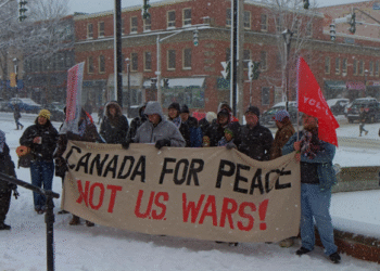 A group of people in winter coats stand in a snowy downtown square in Fredericton, holding a large banner reading “Canada for Peace Not U.S. wars!” Snow falls heavily and brick buildings line the street behind them.
