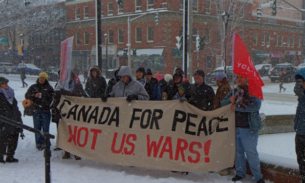 A group of people in winter coats stand in a snowy downtown square in Fredericton, holding a large banner reading “Canada for Peace Not U.S. wars!” Snow falls heavily and brick buildings line the street behind them.