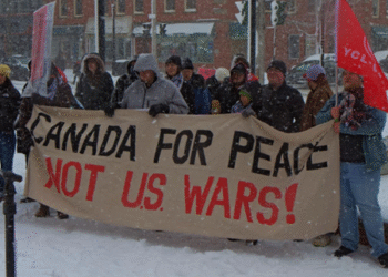 A group of people in winter coats stand in a snowy downtown square in Fredericton, holding a large banner reading “Canada for Peace Not U.S. wars!” Snow falls heavily and brick buildings line the street behind them.