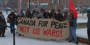 A group of people in winter coats stand in a snowy downtown square in Fredericton, holding a large banner reading “Canada for Peace Not U.S. wars!” Snow falls heavily and brick buildings line the street behind them.