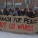 A group of people in winter coats stand in a snowy downtown square in Fredericton, holding a large banner reading “Canada for Peace Not U.S. wars!” Snow falls heavily and brick buildings line the street behind them.