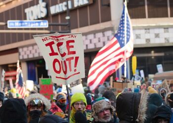 Crowd of protesters in winter clothing gathered in downtown Minneapolis holding “ICE Out” signs and U.S. flags during a demonstration against Immigration and Customs Enforcement.