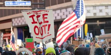 Crowd of protesters in winter clothing gathered in downtown Minneapolis holding “ICE Out” signs and U.S. flags during a demonstration against Immigration and Customs Enforcement.
