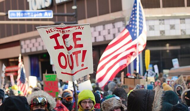 Crowd of protesters in winter clothing gathered in downtown Minneapolis holding “ICE Out” signs and U.S. flags during a demonstration against Immigration and Customs Enforcement.