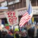 Crowd of protesters in winter clothing gathered in downtown Minneapolis holding “ICE Out” signs and U.S. flags during a demonstration against Immigration and Customs Enforcement.
