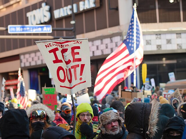 Crowd of protesters in winter clothing gathered in downtown Minneapolis holding “ICE Out” signs and U.S. flags during a demonstration against Immigration and Customs Enforcement.